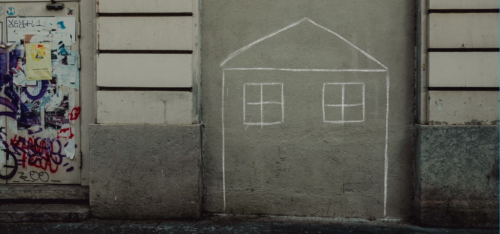 fotografía de un dibujo a tiza  en el que aparece una casa. El dibujo está hecho en la calle sobre una puerta tapiada con cemento